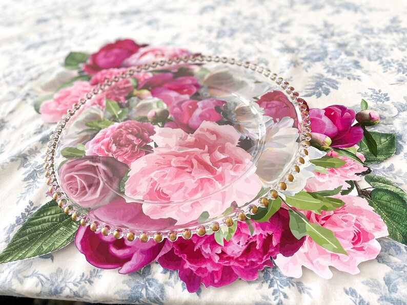 Op de afbeelding: Een heldere glazen serveerschotel met een gouden parelrand ligt op een onderzetter met bloemenprint. De onderzetter heeft roze, witte en groene pioenen.