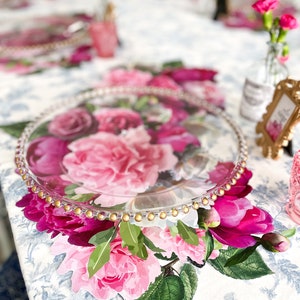Op de afbeelding: Een doorzichtig glazen bord met een gouden parelrand en een bloemenontwerp met roze en paarse rozen. Het bord ligt op een witte tafelkleed met een bloemenpatroon.