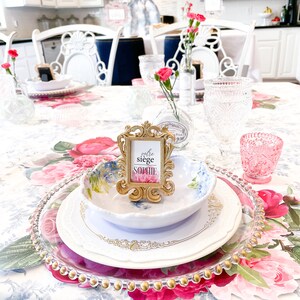 Op de afbeelding: Een witte tafelsetting met een bloemen tafelkleed. Een gouden fotolijst met de tekst "votre si&egrave;ge Sophie" staat op de tafel. Er staan borden, kommen en glazen op de tafel.