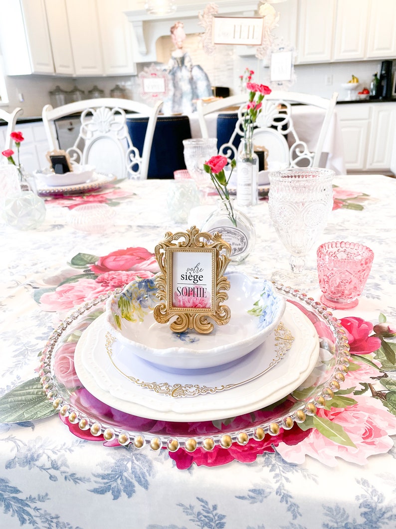 Op de afbeelding: Een witte tafelsetting met een bloemen tafelkleed. Een gouden fotolijst met de tekst "votre si&egrave;ge Sophie" staat op de tafel. Er staan borden, kommen en glazen op de tafel.