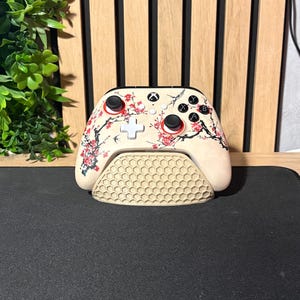 Op de afbeelding: Een beige Xbox-controller met een bloemmotief van kersenbloesem, rustend op een bijpassende standaard. De controller heeft zwarte knoppen en rode accenten. De standaard heeft een honingraatpatroon.