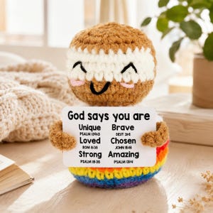 May include: A handmade crochet doll with a smiling face, holding a sign that reads "God says you are Unique, Brave, Loved, Chosen, Strong, Amazing." The doll has a brown head, white and brown striped middle, and a rainbow-colored base.