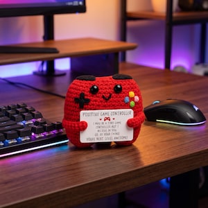 May include: A red crocheted plush toy shaped like a game controller with black eyes and a smile, holding a white card that reads: POSITIVE GAME CONTROLLER, I MAY BE A TINY GAME CONTROLLER BUT I BELIEVE IN YOU, GO DO YOUR THING! YOU'RE NEXT LEVEL AWESOME!