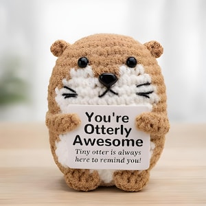 May include: A brown and white crocheted otter plush toy holding a white rectangular sign with black text that reads: You're Otterly Awesome. Tiny otter is always here to remind you! The otter has black plastic eyes and a black nose.
