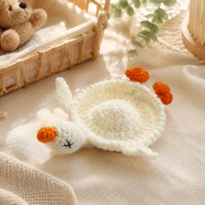 May include: A handmade, off-white crochet goose coaster with orange accents. The goose has a white body, a small orange beak, and two orange feet. The coaster is designed to resemble a goose.