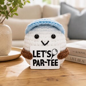 Op de afbeelding: Een wit gehaakt golfbal-personage met een lichtblauwe hoed en bruine armbanden, met een bord met de tekst "LET'S PAR-TEE" met een golfbal- en tee-afbeelding. Het personage heeft zwarte ogen en een lachend gezicht.