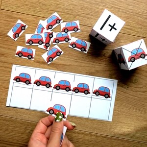 May include: Educational counting game featuring red car cutouts, a number line, and dice with math symbols. The cars have blue windows and black tires. The game is designed for early learning and number recognition.
