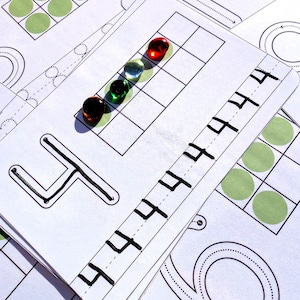 May include: White educational cards with black outlines and the number 4 printed on them. Five colourful marbles, including red, green, and clear, are arranged on one card. Green circles are also printed on the cards.