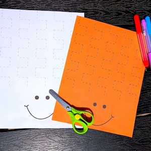 May include: Two sheets of paper, one white and one orange, with a dotted line pattern and a smiling face drawn on them. A pair of green-handled scissors and three colored markers are also present.