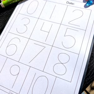 May include: A white worksheet with dotted lines forming the numbers 0 through 10. The worksheet is in a clear plastic sleeve. Colorful markers and decorative glass beads are visible in the background. The worksheet is for educational purposes.