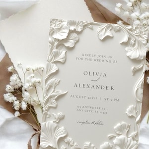 May include: A rectangular wedding invitation card with a cream-colored background and raised white floral relief borders. The text reads: Kindly join us for the wedding of Olivia and Alexander, August 20th at 5 PM, 123 Anywhere St., Any City, ST 12345, reception to follow.