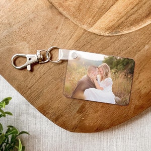 May include: Rectangular metal keychain with a photo of a couple embracing in a field. The keychain has a silver clasp and a clear plastic strap. The photo shows a man and woman in a natural setting.