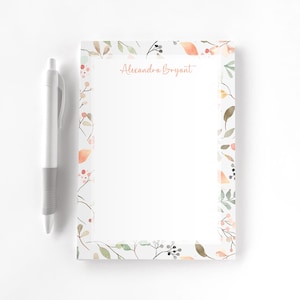 May include: A white notepad with a floral border featuring watercolor leaves and berries in shades of green, orange, and pink. The notepad has the name "Alexandra Bryant" printed in a delicate script. A silver and gray pen rests beside the notepad.