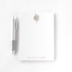 May include: A white notepad with a pink cross decorated with pink and green flowers. The notepad has the name "Christine Smith" printed at the bottom.