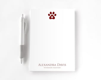 May include: A white notepad featuring a dark red paw print icon with a small white cross in the center at the top, and the text "ALEXANDRA DAVIS" above "VETERINARY ASSISTANT" in dark red at the bottom, accompanied by a white and gray pen.