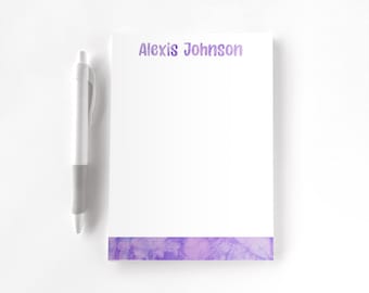 May include: A white notepad featuring the name Alexis Johnson printed in purple at the top and a purple watercolour-style tie-dye pattern at the bottom, accompanied by a white and grey retractable ballpoint pen positioned to the left.
