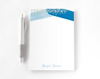 May include: A white notepad featuring a top border of light blue and dark blue brushstrokes with gold glitter accents, and the name Morgan Stevens printed in light blue at the bottom, accompanied by a white and grey retractable ballpoint pen.