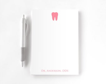 May include: A white notepad featuring a pink tooth icon at the top and the text DR. ANDERSON, DDS in pink at the bottom, accompanied by a white and gray retractable ballpoint pen positioned to the left of the notepad.