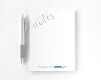 May include: A white notepad featuring the word notes in a grey cursive script at the top left and the name MACKENZIE JOHNSON printed in blue capital letters at the bottom, accompanied by a white and grey retractable ballpoint pen.