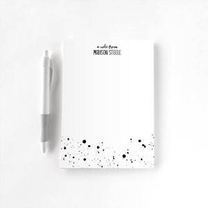 May include: A white notepad with black ink splatter at the bottom. The notepad has the text "a note from MADISON STEELE" at the top. A white pen is next to the notepad.
