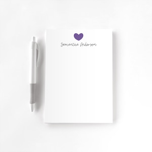 May include: A white notepad featuring a purple heart icon and the printed name Samantha Anderson in black script, accompanied by a white and gray retractable ballpoint pen positioned to the left of the notepad.