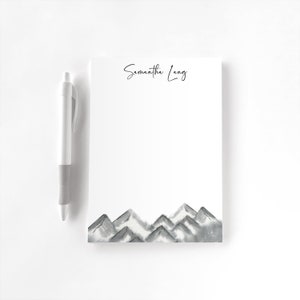 May include: A white notepad with a watercolor mountain range design at the bottom and the name "Samantha Lang" in script at the top. A white and gray pen rests on the left side of the notepad. The notepad is blank.