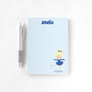 May include: A pale blue notepad featuring the name "Amelia" in dark blue. A cartoon ballerina in a blue dress and white ice skates is depicted. A white pen with a grey grip is placed beside the notepad.