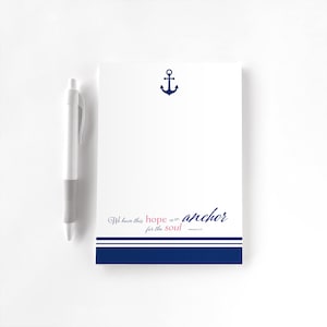 May include: White notepad with a navy blue anchor graphic and the text "We have this hope as an anchor for the soul" in pink and navy blue. A white pen with a grey grip is on the left.