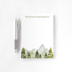 May include: A white notepad with a watercolor forest and mountain design. The notepad has the name "NICHOLAS BLACKSMITH" printed at the top. A white and gray pen rests beside the notepad.