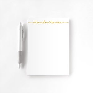 Peut inclure: Un bloc-notes blanc avec une inscription en or cursive qui dit "alexandra stevenson" en haut. Un stylo blanc avec une poignée grise est placé à gauche du bloc-notes.