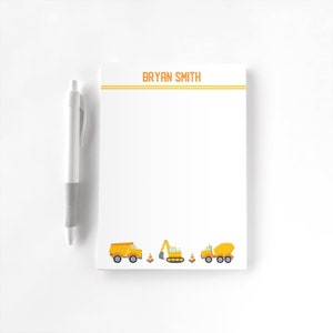 May include: White notepad with the name "BRYAN SMITH" in orange letters at the top. The notepad has a construction theme with illustrations of a dump truck, excavator, and cement mixer. A white pen with a grey grip is on the left.