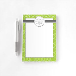 May include: A notepad with a white interior and a lime green border featuring a white floral pattern. The notepad has a grey horizontal stripe and a circular emblem with the text "Be Still and Know That I Am God". A white pen with a grey grip is to the left.