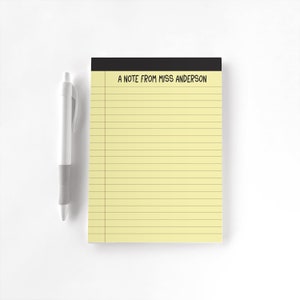 May include: A yellow notepad with horizontal lines and a black header featuring the text A NOTE FROM MISS ANDERSON in black capital letters, accompanied by a white retractable ballpoint pen with a gray rubber grip section.