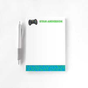 May include: A white notepad with a green gamer controller graphic and the text "RYAN ANDERSON" at the top. The notepad has a blue and green border at the bottom.