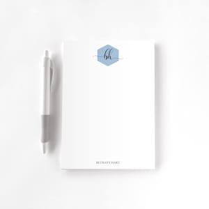 May include: A white notepad with a light blue hexagon design containing the letters "bh" in black script. A white pen with a gray grip rests beside the notepad. The name "BETHANY HART" is printed at the bottom of the notepad.