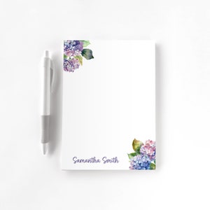 May include: White notepad with watercolor hydrangea floral design in purple and blue. The notepad has the name "Samantha Smith" printed in a script font. A white pen is placed next to the notepad.