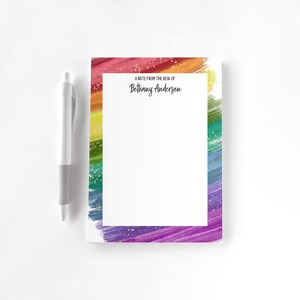 May include: A notepad with a rainbow-coloured brushstroke border and gold flecks. The notepad has the text "A note from the desk of Bethany Anderson" and a white pen.