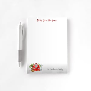 May include: A white notepad with the text "Notes from the farm" in red script. The bottom of the notepad features a watercolor illustration of a red barn, a windmill, and the text "The Sanderson Family". A white pen rests on the left.