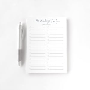 May include: A white notepad with a grocery list layout and the text "the shrombergh family GROCERY LIST" in navy blue script. A white and gray pen rests beside the notepad. The notepad has lines and check boxes.