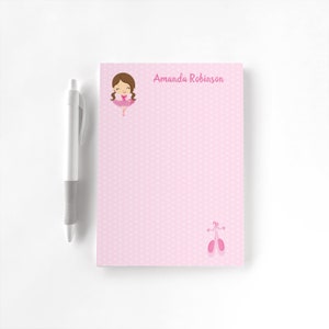 May include: Pink notepad with a polka dot pattern, featuring a cartoon ballerina illustration and the name "Amanda Robinson" in pink text. A silver and gray pen is next to the notepad. Ballet shoes are at the bottom.