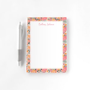 May include: A floral notepad with a white background and a border of pink, orange, and green flowers. The notepad has the name "Brittany Anderson" printed in pink. A white and gray pen is placed to the left of the notepad.