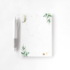 May include: A white notepad with a marble design, gold accents, and green watercolor leaves. A gold letter "J" is in the upper left corner. A white and gray pen is to the left of the notepad.