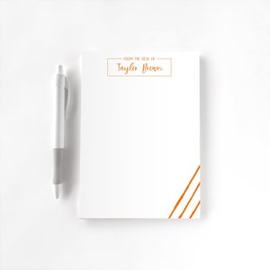 May include: A white notepad with the text "FROM THE DESK OF Taylor Brown" in a gold-orange color. Three diagonal lines in the same color are in the lower right corner. A white and gray pen is to the left of the notepad.