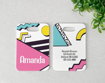 May include: Two rectangular luggage tags with rounded corners and a top slot. The left tag features a pink triangle, black stripes, a yellow circle, and the name Amanda. The right tag displays contact information for Amanda Stevens with geometric shapes.