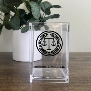 May include: A clear acrylic desk organizer with a black emblem of scales of justice inside a laurel wreath. The text "Anthony Bryant, J.D." is printed below the emblem. A white potted plant is in the background.