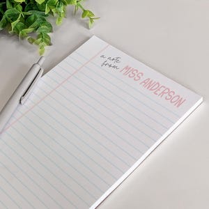 Puede incluir: Un bloc de notas blanco con l&iacute;neas azules y rosas y el texto "a note from MISS ANDERSON" en rosa y negro. Un bol&iacute;grafo plateado descansa sobre el bloc de notas. Planta de hojas verdes en el fondo.