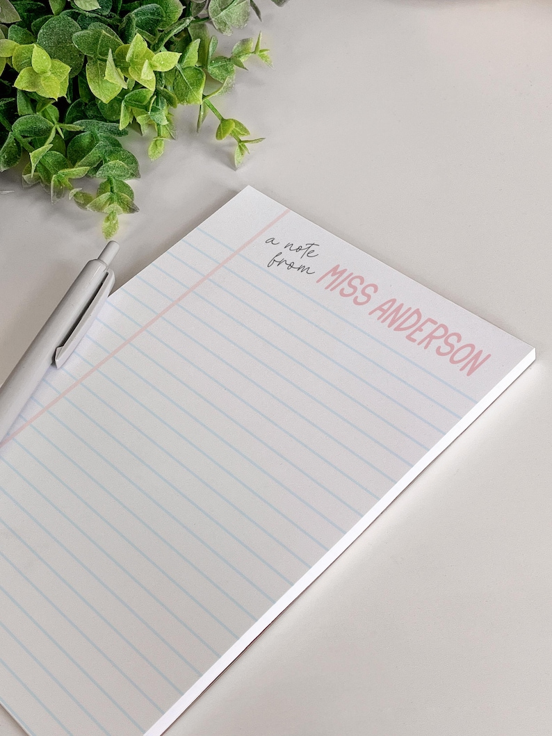 Puede incluir: Un bloc de notas blanco con l&iacute;neas azules y rosas y el texto "a note from MISS ANDERSON" en rosa y negro. Un bol&iacute;grafo plateado descansa sobre el bloc de notas. Planta de hojas verdes en el fondo.