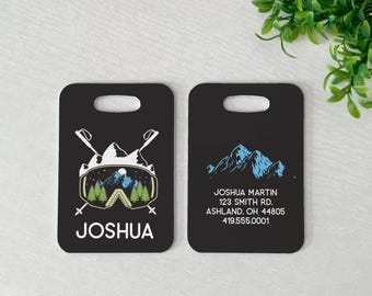 May include: Two black rectangular luggage tags with rounded corners and a top handle slot. The left tag features a ski goggle graphic with mountains, trees, and the name JOSHUA. The right tag displays a mountain range and contact information.