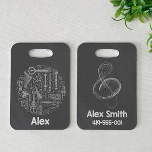 May include: Two dark gray luggage tags. One tag features a white illustration of musical instruments and the name "Alex". The other tag has a treble clef and the name "Alex Smith" with a phone number.