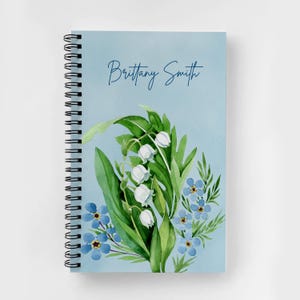 May include: A spiral-bound notebook with a light blue cover featuring a watercolor illustration of white lily of the valley flowers and small blue forget-me-not flowers with green leaves. The name Brittany Smith is printed in dark blue script.